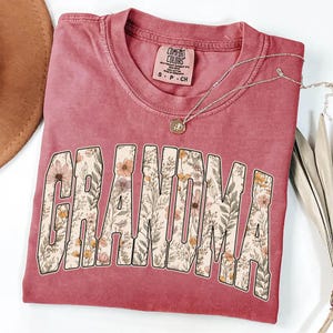 Op de afbeelding: Een stoffig roze Comfort Colors T-shirt met het woord "GRANDMA" in een bloemenontwerp. Een zilveren ketting met een hanger ligt op het gevouwen shirt. Het shirt is nieuw.