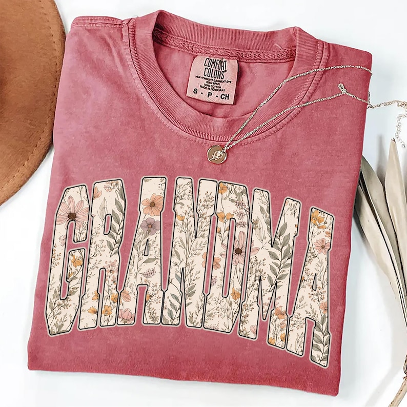 Op de afbeelding: Een stoffig roze Comfort Colors T-shirt met het woord "GRANDMA" in een bloemenontwerp. Een zilveren ketting met een hanger ligt op het gevouwen shirt. Het shirt is nieuw.