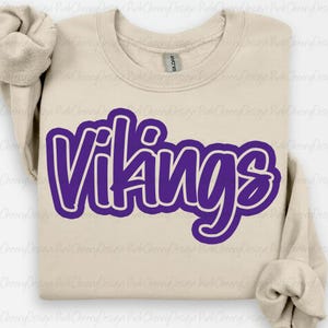 Può includere: Una felpa beige con la scritta "Vikings" in grandi lettere viola a bolle. La felpa ha un girocollo e maniche lunghe. L'etichetta del marchio Gildan è visibile all'interno del colletto.