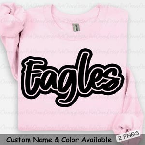 May include: A light pink sweatshirt with the word "Eagles" in large, black, cursive lettering outlined in white. The text "Custom Name & Color Available" is at the bottom. The sweatshirt is folded, with the sleeves visible.