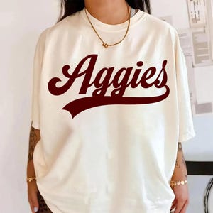 May include: Cream-coloured t-shirt featuring the word "Aggies" in maroon script. The shirt has short sleeves and a relaxed fit. A gold necklace is visible at the neckline.