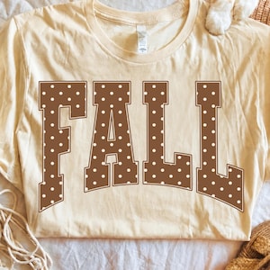 May include: A cream-colored short-sleeve t-shirt featuring the word FALL printed in large, brown, block-style letters. Each letter is decorated with a repeating pattern of small white polka dots. The shirt has a crew neckline and a standard fit.