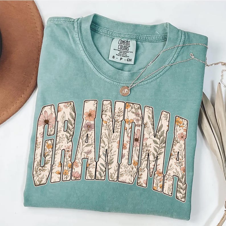 Op de afbeelding: Een zeegroen t-shirt met het woord "GRANDMA" in grote, bloemrijke letters. Het shirt heeft een ronde hals en korte mouwen. Een zilveren ketting met een hanger is over het shirt gedrapeerd.