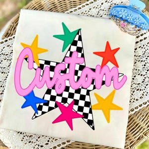 May include: A white t-shirt featuring a large central star with a black and white checkered pattern, surrounded by smaller stars in yellow, teal, red, blue, and pink. The word Custom is printed across the center in a large, pink, cursive font.
