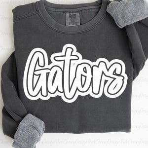 May include: A dark gray sweatshirt with the word "Gators" in large white, cursive lettering. The sweatshirt has a crew neck and long sleeves with a gray, fuzzy lining visible at the cuffs. The text is outlined in black.