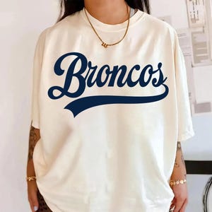 Puede incluir: Camiseta color crema con la palabra "Broncos" en escritura azul marino en la parte delantera. La palabra está arqueada y tiene una línea azul marino debajo. La camiseta tiene mangas cortas y un corte holgado.