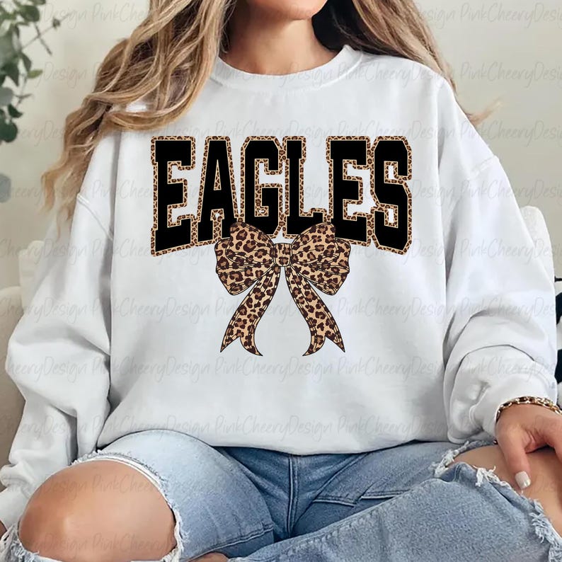 May include: White sweatshirt with the word "EAGLES" in black block letters with a leopard print fill. A leopard print bow is centered below the word. The sweatshirt is a casual style.