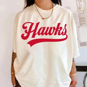 May include: Cream-colored t-shirt with the word "Hawks" in red script across the front. The word is arched above a red banner. The shirt has short sleeves and a relaxed fit.