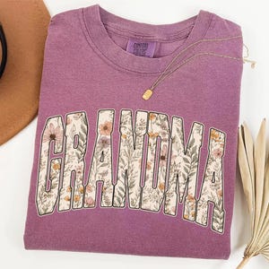Op de afbeelding: Een mauvekleurige sweatshirt met het woord "GRANDMA" in een bloemenmotief. De letters zijn gevuld met een patroon van bloemen en bladeren in roze, gele en groene tinten. Een gouden ketting met een kleine hanger ligt over het shirt.