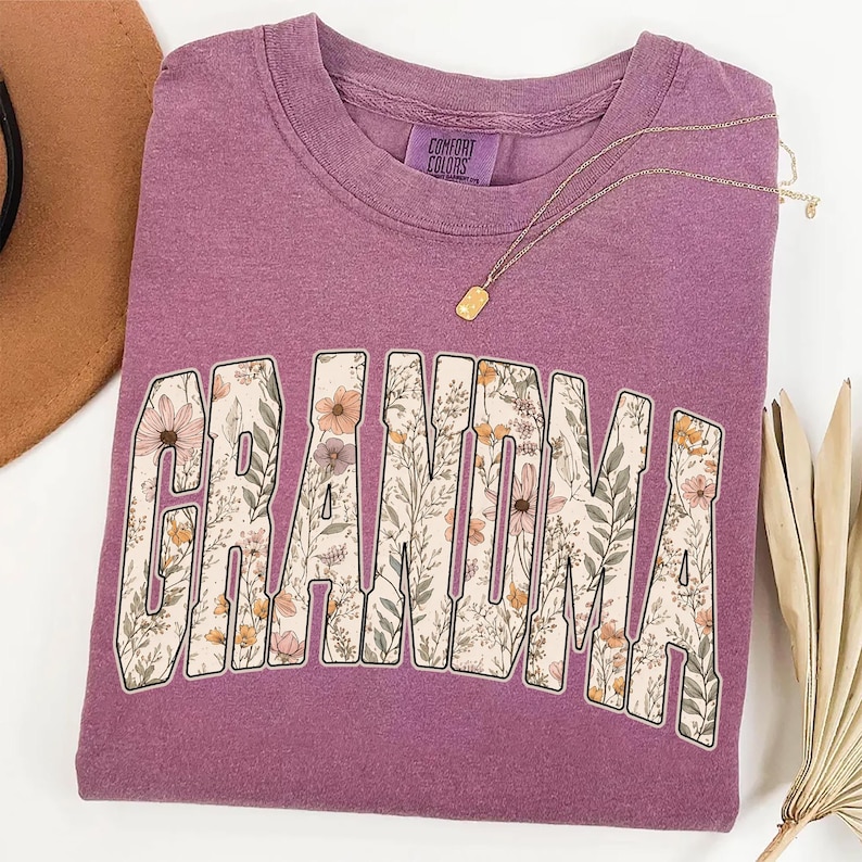 Op de afbeelding: Een mauvekleurige sweatshirt met het woord "GRANDMA" in een bloemenmotief. De letters zijn gevuld met een patroon van bloemen en bladeren in roze, gele en groene tinten. Een gouden ketting met een kleine hanger ligt over het shirt.