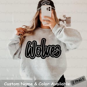 May include: A light gray sweatshirt with the word "Wolves" in a black and white outlined font. The text "Custom Name & Color Available" is at the bottom. The image also includes a black baseball cap.