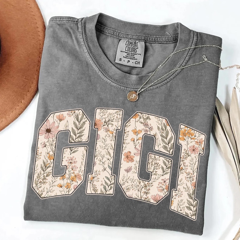 May include: A gray Comfort Colors t-shirt with the word "GIGI" in large, floral-patterned letters. A silver necklace with a small pendant rests on the shirt. The shirt is folded, and a brown hat is partially visible.