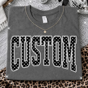Pode incluir: Uma t-shirt de gola redonda cinzento-carvão com a palavra CUSTOM impressa em letras pretas grandes e negrito, um padrão de bolinhas brancas e um contorno festonado branco estilo renda. Um colar de corrente fina dourada com um pingente de sorriso.