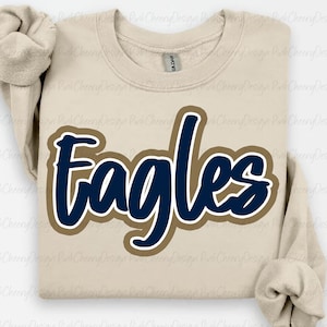 May include: A beige sweatshirt with the word "Eagles" in navy blue script, outlined in a tan color. The sweatshirt has a crew neck and long sleeves. The Gildan brand tag is visible.