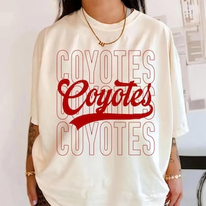May include: Cream-colored t-shirt with the word "COYOTES" in a stacked design, with the word "Coyotes" in a cursive font in the middle. The text is in red. The shirt has short sleeves.