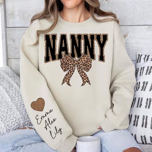 Puede incluir: Una sudadera color crema con la palabra "NANNY" en letras negras con relleno de estampado de leopardo. Un lazo con estampado de leopardo est&aacute; centrado debajo de la palabra. La manga izquierda tiene un coraz&oacute;n con estampado de leopardo y los nombres "Emma, Alex, Lily" en cursiva.