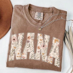 May include: Brown Comfort Colors t-shirt with the word "NANA" in large, floral-filled letters. The shirt has a crew neck and short sleeves. A silver necklace with a small pendant is draped across the shirt.
