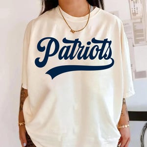 Pode incluir: Camiseta creme com a palavra "Patriots" em escrita azul marinho e uma faixa azul marinho por baixo. A camiseta tem mangas curtas e um corte folgado, adequada para uso casual.