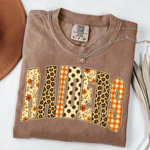 May include: A brown short-sleeved t-shirt with a crew neckline and a Comfort Colors brand label. The front features the word RAIDERS in large block letters, each filled with a different pattern including orange floral, leopard print, and orange plaid.