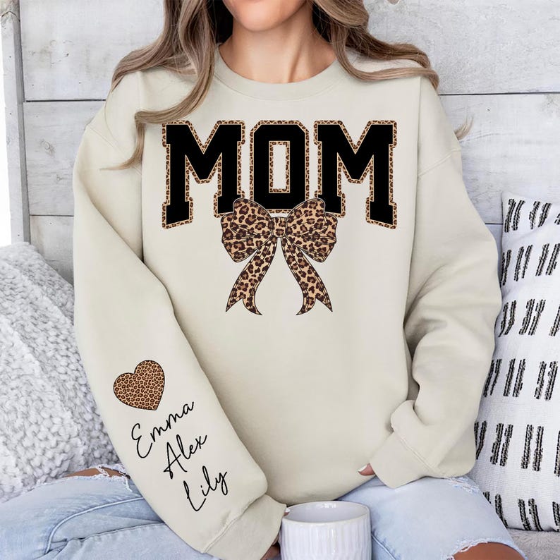 Puede incluir: Una sudadera color crema con la palabra "MOM" en letras negras con estampado de leopardo. Un lazo con estampado de leopardo est&aacute; centrado debajo de la palabra. La manga izquierda presenta un coraz&oacute;n con estampado de leopardo y los nombres "Emma, Alex, Lily."