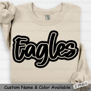 May include: A beige crewneck sweatshirt with the word "Eagles" in large, black, outlined lettering across the front. The text "Custom Name & Color Available" is at the bottom. The sleeves are folded.