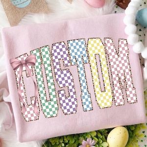 Puede incluir: Sudadera rosa claro con la palabra "CUSTOM" en letras grandes y coloridas a cuadros. Las letras están delineadas con un estampado de leopardo. Un lazo rosa está en el lado izquierdo de la palabra.