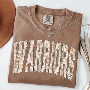 Puede incluir: Una camiseta marr&oacute;n con la palabra "WARRIORS" en letras grandes y florales. La camiseta tiene cuello redondo y mangas cortas. Un collar plateado con un peque&ntilde;o colgante est&aacute; sobre la camiseta.