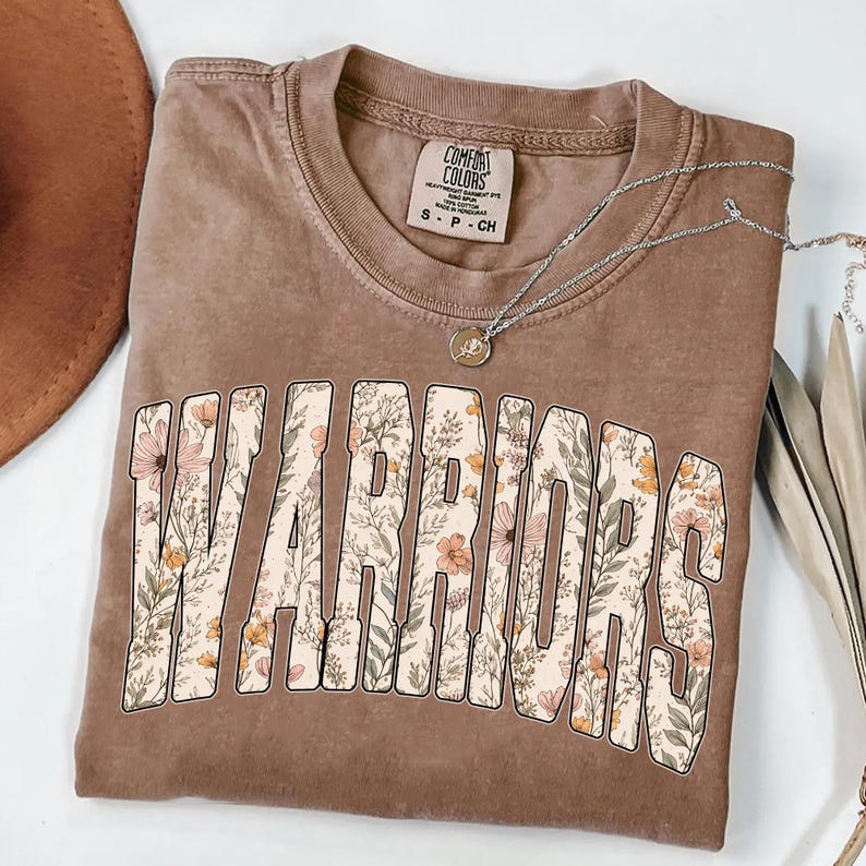 Puede incluir: Una camiseta marr&oacute;n con la palabra "WARRIORS" en letras grandes y florales. La camiseta tiene cuello redondo y mangas cortas. Un collar plateado con un peque&ntilde;o colgante est&aacute; sobre la camiseta.