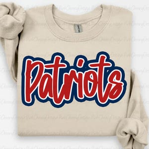Pode incluir: Camisola bege com a palavra "Patriots" em vermelho, contornada em azul e branco. A camisola tem mangas compridas e gola redonda. A etiqueta da marca Gildan é visível na gola.