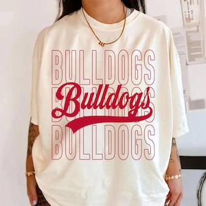 May include: Cream-colored t-shirt with the word "BULLDOGS" in red, repeated three times. The middle word is in a script font with a red swoosh underneath. The shirt has short sleeves and a relaxed fit.