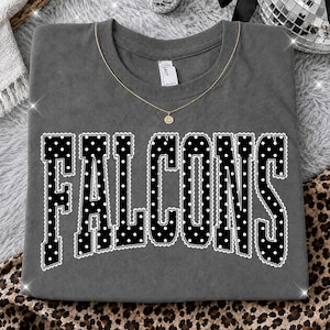 Puede incluir: Una camiseta de manga corta gris carbón que presenta la palabra FALCONS en letras mayúsculas grandes y negras con un patrón de lunares blancos y un contorno festoneado blanco. Una fina cadena dorada con un dije de carita feliz descansa en el cuello.