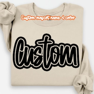 May include: A beige sweatshirt with the word "Custom" in large, black, outlined lettering. Above the word "Custom" is the text "Custom mascot name & color" in orange. The sleeves are folded.
