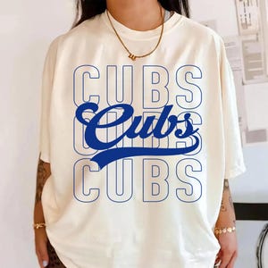 Puede incluir: Camiseta de color crema con la palabra "CUBS" en un diseño azul en capas. La palabra "Cubs" se repite tres veces, la del medio en cursiva. La camiseta tiene mangas cortas y cuello redondo.