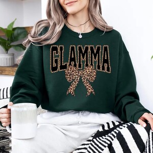 May include: A dark green sweatshirt with the word "GLAMMA" in black block letters outlined in gold. A leopard print bow is centered below the text. The person is holding a white mug.
