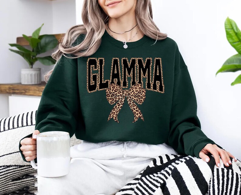 May include: A dark green sweatshirt with the word "GLAMMA" in black block letters outlined in gold. A leopard print bow is centered below the text. The person is holding a white mug.