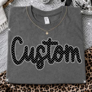 Peut inclure: Un t-shirt à col rond gris anthracite avec le mot Custom écrit en lettres cursives noires à pois blancs et contour blanc. Un fin collier à chaîne dorée avec un petit pendentif en forme de visage souriant repose sur l'encolure.