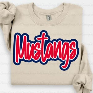May include: A beige Gildan sweatshirt featuring the word "Mustangs" in red, outlined in blue. The word is in a bold, cursive font. The sweatshirt has long sleeves and a crew neck.