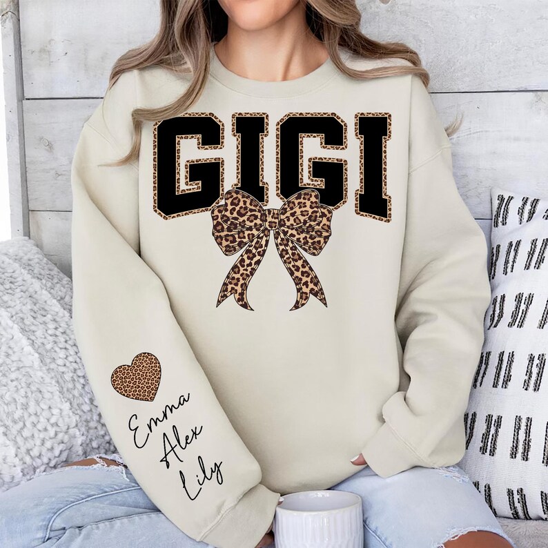 Puede incluir: Sudadera color crema con la palabra "GIGI" en letras negras con relleno y ribete de estampado de leopardo. Un lazo con estampado de leopardo est&aacute; centrado debajo del texto. Un coraz&oacute;n con estampado de leopardo y nombres est&aacute;n en la manga izquierda.