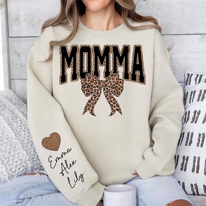 Puede incluir: Sudadera color crema con la palabra "MOMMA" en letras negras con estampado de leopardo. Un lazo con estampado de leopardo est&aacute; debajo de la palabra. La manga izquierda tiene un coraz&oacute;n con estampado de leopardo y los nombres "Emma", "Alex" y "Lily".