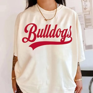 K&ouml;nnte beinhalten: Cremefarbenes T-Shirt mit dem Wort "Bulldogs" in roter Schrift auf der Vorderseite. Das Wort ist &uuml;ber einem roten Banner gew&ouml;lbt. Das Shirt hat kurze &Auml;rmel und eine lockere Passform.