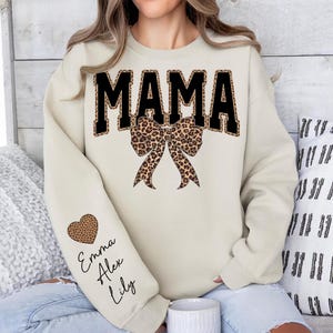 Puede incluir: Sudadera color crema con la palabra "MAMA" en letras negras grandes con estampado de leopardo. Un lazo con estampado de leopardo est&aacute; debajo de la palabra. La manga izquierda tiene un coraz&oacute;n con estampado de leopardo con los nombres "Emma", "Alex" y "Lily".