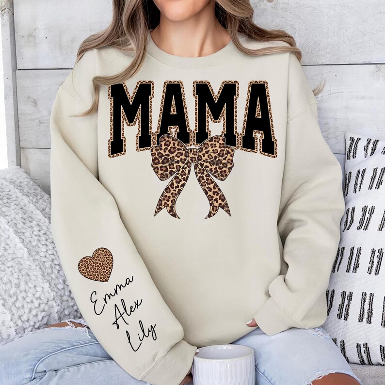 Puede incluir: Sudadera color crema con la palabra "MAMA" en letras negras grandes con estampado de leopardo. Un lazo con estampado de leopardo est&aacute; debajo de la palabra. La manga izquierda tiene un coraz&oacute;n con estampado de leopardo con los nombres "Emma", "Alex" y "Lily".