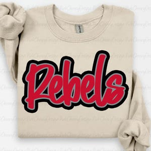 May include: A beige sweatshirt with the word "Rebels" in red, outlined in black and gray. The sweatshirt has a crew neck and long sleeves. The Gildan brand tag is visible on the inside of the collar.