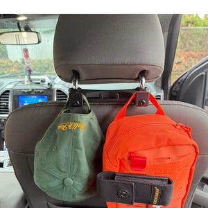 May include: A car headrest organizer with two black hooks holding a green baseball cap and an orange pouch. A black Leatherman tool holder is attached to the orange pouch. The baseball cap has yellow text.