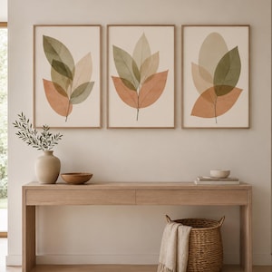 Puede incluir: Tres impresiones artísticas enmarcadas con diseños abstractos de hojas en tonos verdes, melocotón y beige. Se exhiben sobre una consola de madera con elementos decorativos, como un jarrón, un cuenco y una cesta tejida. La habitación tiene una paleta de colores neutros.