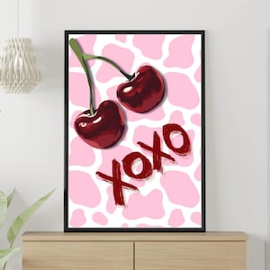 Puede incluir: Una impresión artística enmarcada con dos cerezas maduras con tallos verdes sobre un fondo rosa y blanco con estampado de vaca. La palabra "xoxo" está escrita en rojo debajo. La obra de arte se exhibe en una cómoda de madera.