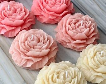 May include: A collection of flower-shaped candles in various shades of pink and cream. The candles are intricately designed to resemble blooming peonies, each with a central wick. The arrangement includes candles in shades of light pink, medium pink, and cream.