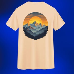 Puede incluir: Una camiseta de color beige claro con un gráfico circular de una cordillera al atardecer. El diseño incluye un sol naranja brillante, montañas azules y grises y árboles de color verde oscuro. La camiseta está hecha de un material suave.
