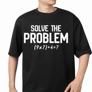 Puede incluir: Camiseta negra con el texto "SOLVE THE PROBLEM" en letras mayúsculas blancas. Debajo, la ecuación matemática (9x7)+4=? también en blanco. La camiseta tiene mangas cortas y cuello redondo.