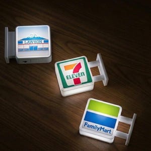May include: Three illuminated square signs with the logos of Lawson, 7-Eleven, and FamilyMart. Each sign is white with a colored logo and a mounting bracket. The Lawson sign features a mountain scene. The signs are on a dark wood surface.